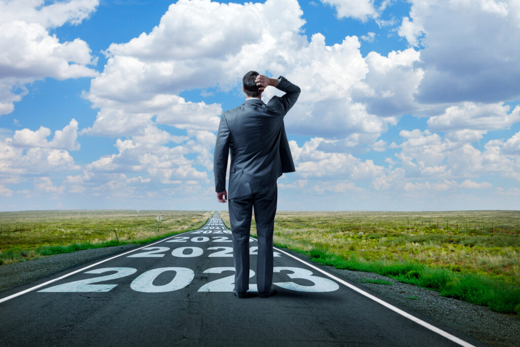 businessman stands on and looks down a long, straight, rural road that has "2023" and subsequent future years painted on it. In the distance, as the road meets the horizon, puffy clouds punctuate the blue sky.
