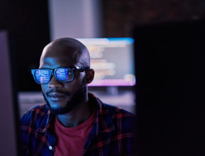 Cyber security, black man and code reflection in eyewear, hacking and software update in office. African American male employee, programmer or IT specialist with glasses, focus and cloud computing